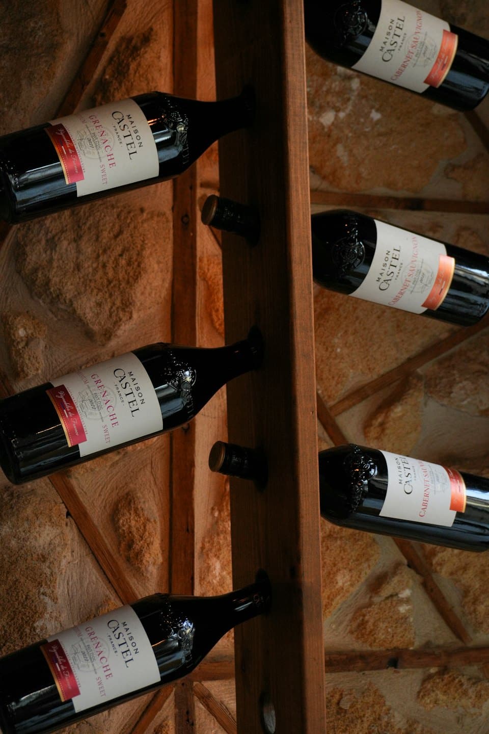 Bottiglie di vino scuro appoggiate orizzontalmente su una rastrelliera di legno contro un muro di pietra.