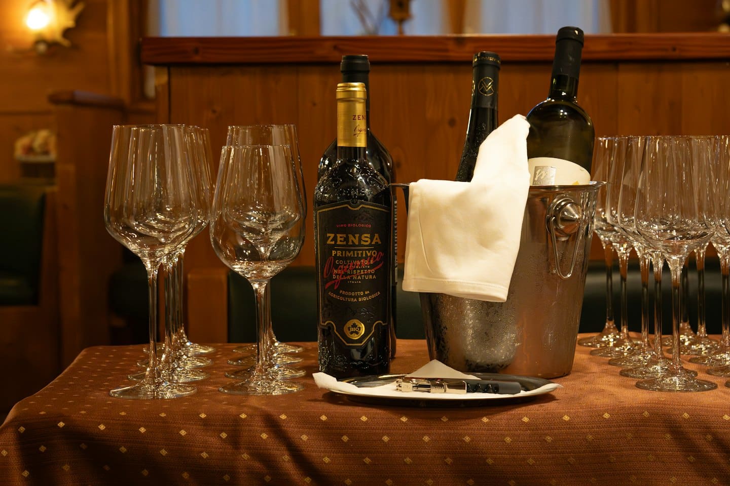 Tavola apparecchiata con bicchieri da vino, bottiglie e secchiello per il ghiaccio.